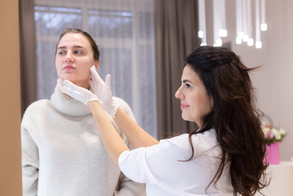 Two women, a beautician doctor and a client, stand at the mirror, at a consultation, discussing the upcoming procedures. beautician talks about sculpting the face